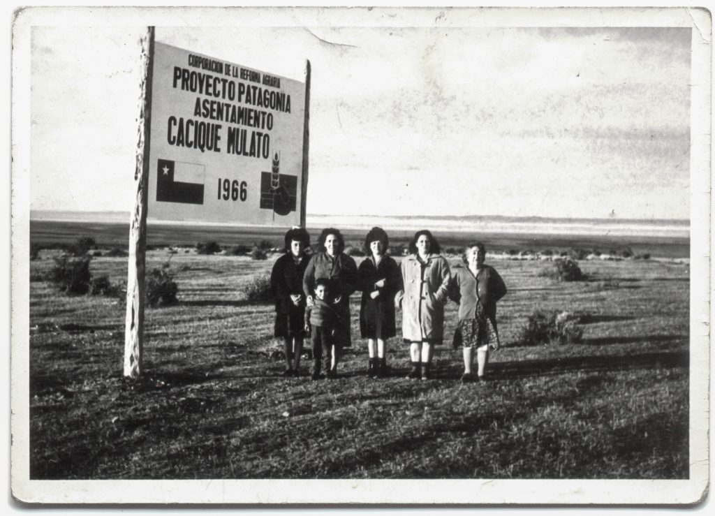 Fotografia que muestra a 5 mujeres pobladoras y un niño al lado de un cartel que se encuentran 
a las afueras del acceso de la cooperativa ¨Cacique Mulato¨. El cartel contiene la frase 
¨Corporación de la reforma agraria, proyecto patagonia asentamiento cacique mulato, 1966¨.